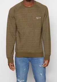 Olijfgroene sweatshirt met een gestructureerd breipatroon, ronde hals, geribde manchetten en een klein wit "Originals" logo op de borst. Draagt met lichtblauwe versleten jeans.