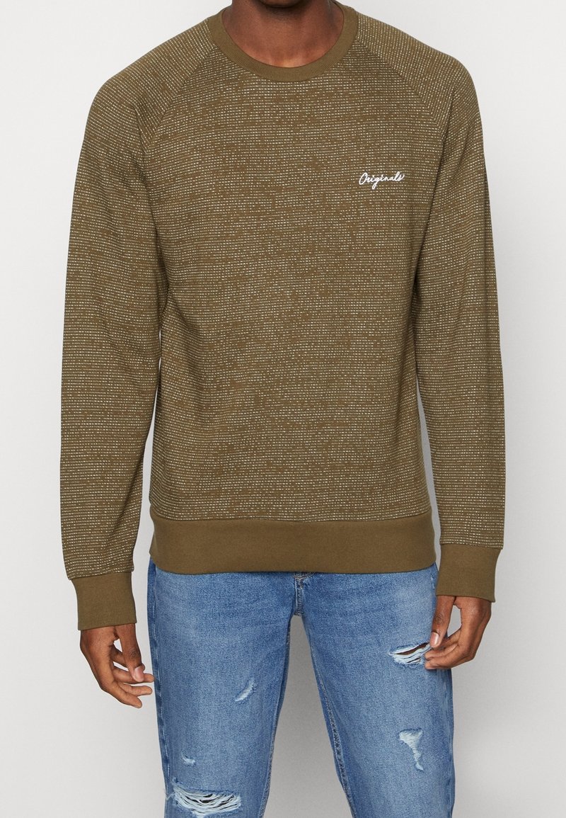 Olijfgroene sweatshirt met een gestructureerd breipatroon, ronde hals, geribde manchetten en een klein wit "Originals" logo op de borst. Draagt met lichtblauwe versleten jeans.