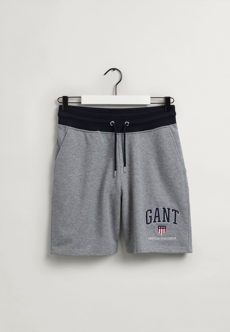 Shorts en coton gris avec ceinture élastique bleu marine et cordons, poches latérales, et logo "GANT American Sportswear" sur la jambe gauche, suspendus sur un cintre blanc.