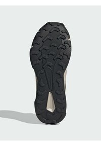 Black rubber outsole with textured pattern and deep treads for traction. Upper features a beige synthetic material and streamlined design.