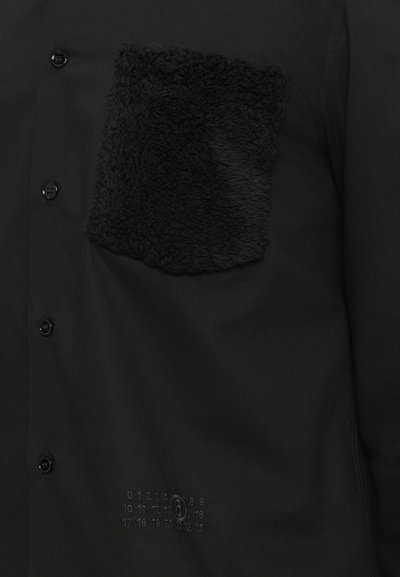 Chemise noire avec une poche en polaire texturée noire, fermeture à boutons et un détail de numérotation en relief sur le bas devant.