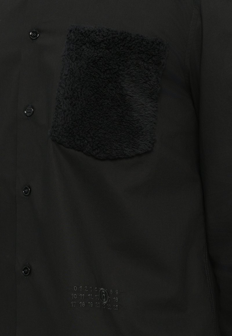 Sort skjorte med en tekstureret sort fleece-lomme, knaplukning og præget nummerdetalje på den nederste front.