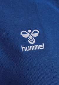 Tissu bleu à texture fine avec un logo brodé blanc d'une abeille stylisée et le mot "hummel" en dessous.