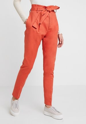 Pantalon orange slim taille haute avec ceinture nouée, porté avec des baskets à plateforme beige et un haut à manches longues côtelé crème.
