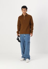 Brauner gerippter Pullover mit einem Reißverschlusskragen, kombiniert mit hellblauen Männer-Cargo-Jeans mit Taschen und weißen Sneakers. Hält einen schwarzen Controller.
