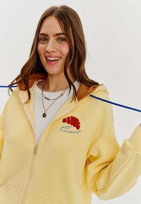 Žltá mikina na zips s hnedou kapucňou, na ktorej je grafika červenej croissant a text "petit croissant". Materiál s bavlneným zložením, s vreckami.