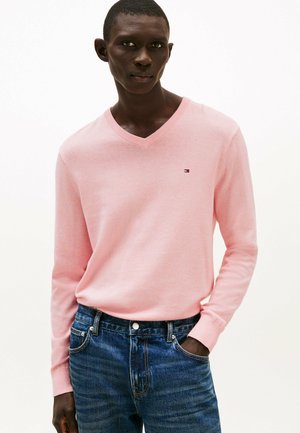 Mand iført en lyserød V-hals sweater med et lille logo på brystet og blå jeans, stående mod en ensfarvet hvid baggrund.