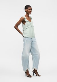 Femme portant un chemisier sans manches vert clair à volants, un jean large déchiré bleu clair et des sandales noires à talons hauts, debout de profil.