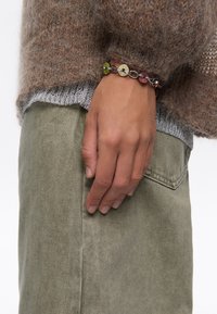 Suéter marrón suave con ribete plateado, pantalones verdes oliva y una pulsera de cuentas multicolores con charms circulares y una cadena metálica.