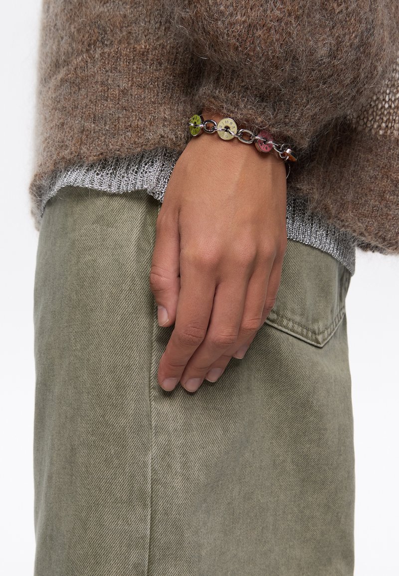 Suéter marrón suave con ribete plateado, pantalones verdes oliva y una pulsera de cuentas multicolores con charms circulares y una cadena metálica.