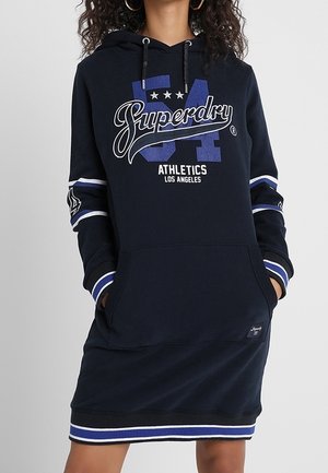 Femme portant une robe à capuche Superdry bleu marine avec des poignets et un ourlet rayés blanc et bleu, mains dans la poche avant.
