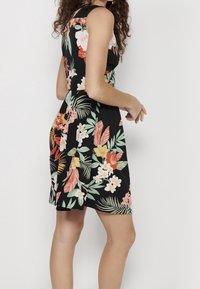 Mujer vistiendo un vestido negro sin mangas con un colorido estampado floral tropical, de pie contra un fondo blanco liso, mirando de lado.