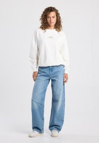 White sweatshirt with a small green logo, paired with wide-leg light blue jeans and white sneakers. Curly hair framing the face.