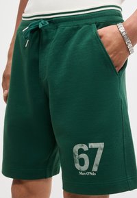 Άνδρας που φοράει πράσινα σορτς με λευκή ριγέ μέση, μπροστινές τσέπες και το λογότυπο "67 Marc O'Polo" τυπωμένο στο αριστερό πόδι.