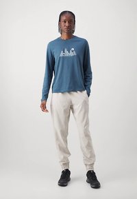 Egy személy áll, kék hosszú ujjú pólót viselve, hegygrafikával, bézs nadrággal és fekete sportcipővel egy egyszerű háttér előtt.