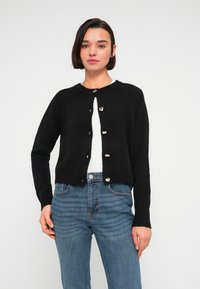 Μαύρη πλεκτή ζακέτα με στρογγυλή λαιμόκοψη, κλείσιμο με κουμπιά και άνετη εφαρμογή, σε συνδυασμό με μπλε τζιν παντελόνι. Λεία υφή, χωρίς σχέδια.