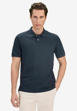 Ung man med kort lockigt hår, klädd i en mörk marinblå pikétröja och ljusbeiga byxor, stående mot en enkel vit bakgrund.