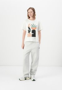 Vrouw in een wit shirt met opdruk, lichte grijze sweatpants en witte sneakers, staand met haar handen in de zakken tegen een effen witte achtergrond.