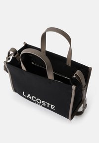 Bolsa de lona negra con asas de cuero gris, que presenta un logo blanco de "LACOSTE" y un compartimento con cremallera en el interior.