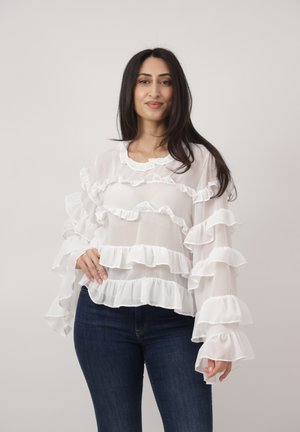 Frau mit langen dunklen Haaren, die eine weiße Bluse mit Rüschen und dunkelblaue Jeans trägt, posiert mit einer Hand in der Hüfte vor neutralem Hintergrund.