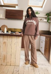 Brauner Sweatshirt mit weißem "ESPRESSO"-Text, karierte braune Hose und beige Hausschuhe, in einer Küche mit Holzakzenten.