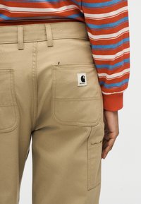 Άτομο που φοράει μπεζ παντελόνια με τσέπες πίσω με πατς, μία από τις οποίες έχει ετικέτα με το λογότυπο Carhartt, και μια μακρυμάνικη μπλούζα με κόκκινες, μπλε και λευκές ρίγες.