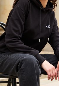 Personne assise sur une chaise, portant un sweat à capuche noir avec un petit logo blanc "CK" et un jean foncé, les mains posées sur les genoux.