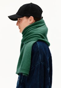 Personne de profil portant une casquette en velours côtelé noire, une veste en velours côtelé bleu marine, et une épaisse écharpe verte enroulée autour du cou.