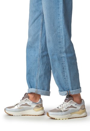 Sneakers caratterizzate da materiali metallici argento e grigi, dettagli in mesh, una suola intermedia bianca e una suola esterna in gomma testurizzata, abbinate a jeans azzurri arrotolati.