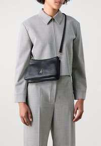 Borsa a tracolla in pelle nera con una texture liscia, caratterizzata da un logo d'insetto dorato e una tracolla rimovibile, indossata su un completo a scacchi bianco e nero.