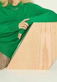 Grönstickad tröja med krage, långa ärmar och en gullänkdetail, i kombination med en texturerad geometrisk träblock.