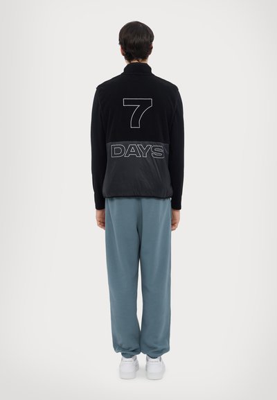 Veste en polaire noire avec "7 DAYS" imprimé en blanc au dos, accompagnée d'un pantalon de survêtement bleu clair et de baskets blanches.