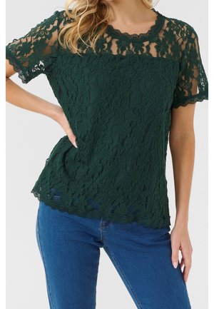 Mujer con blusa de encaje floral en verde oscuro de manga corta combinada con vaqueros azules, de pie con una mano en la cadera contra un fondo claro.