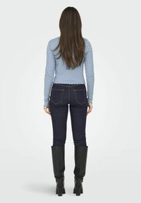 Lysblå langærmet top med ribbet struktur, parret med mørkeblå skinny jeans og knæhøje sorte læderstøvler. Hun står med ryggen til.