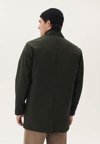 Abrigo impermeable verde oscuro con corte recto, mangas largas y puños abotonados. Presenta una ventilación en la espalda y se complementa con un gorro negro de canalé.