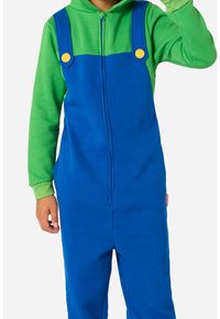 Enfant portant une salopette bleue à boutons jaunes par-dessus un sweat à capuche vert, levant la main droite.
