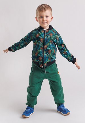 Młody chłopiec uśmiechający się z rozłożonymi ramionami, ubrany w kurtkę z tropikalnym wzorem, zielone spodnie i niebieskie tenisówki, stojący na białym tle.