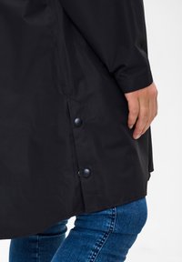 Black waterproof jacket with a smooth texture, featuring a curved hem and two snap buttons on the side. Worn over blue jeans.