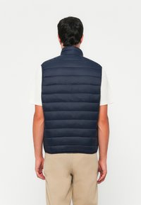 Marineblå polstret vest med høy hals, med horisontale quilte-seksjoner, i kombinasjon med en lysegrå t-skjorte og beige bukser.