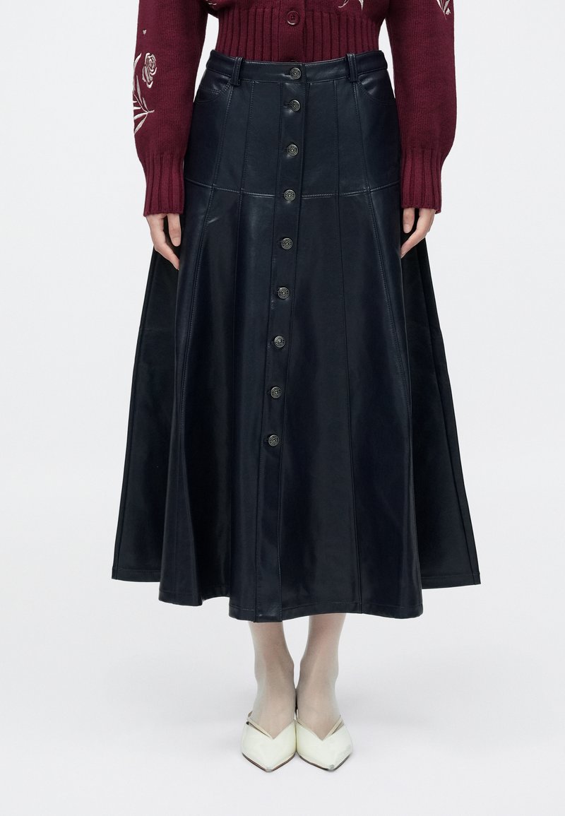 Hosszú fekete bőrszoknya, A-vonalú sziluett, elöl gombos záródással, sima textúrával és bővülő szegéllyel. Fehér, hegyes orrú cipővel párosítva.
