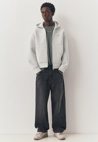 Jeune homme portant un sweat à capuche zippé gris clair, un jean large foncé, une chemise rayée, des baskets beige et un collier ainsi que des bracelets en argent.