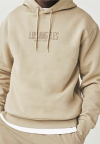 Sudadera beige con capucha ajustable, que presenta "LOS ÁNGELES WEST COAST" en texto marrón claro en el pecho. Textura suave, puños acanalados.