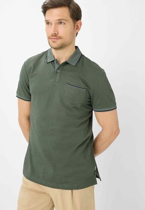 Mann trägt ein dunkelgrünes Polohemd mit kurzen Ärmeln und Brusttasche sowie beige Hose, steht mit den Händen hinter dem Rücken und blickt leicht nach rechts.