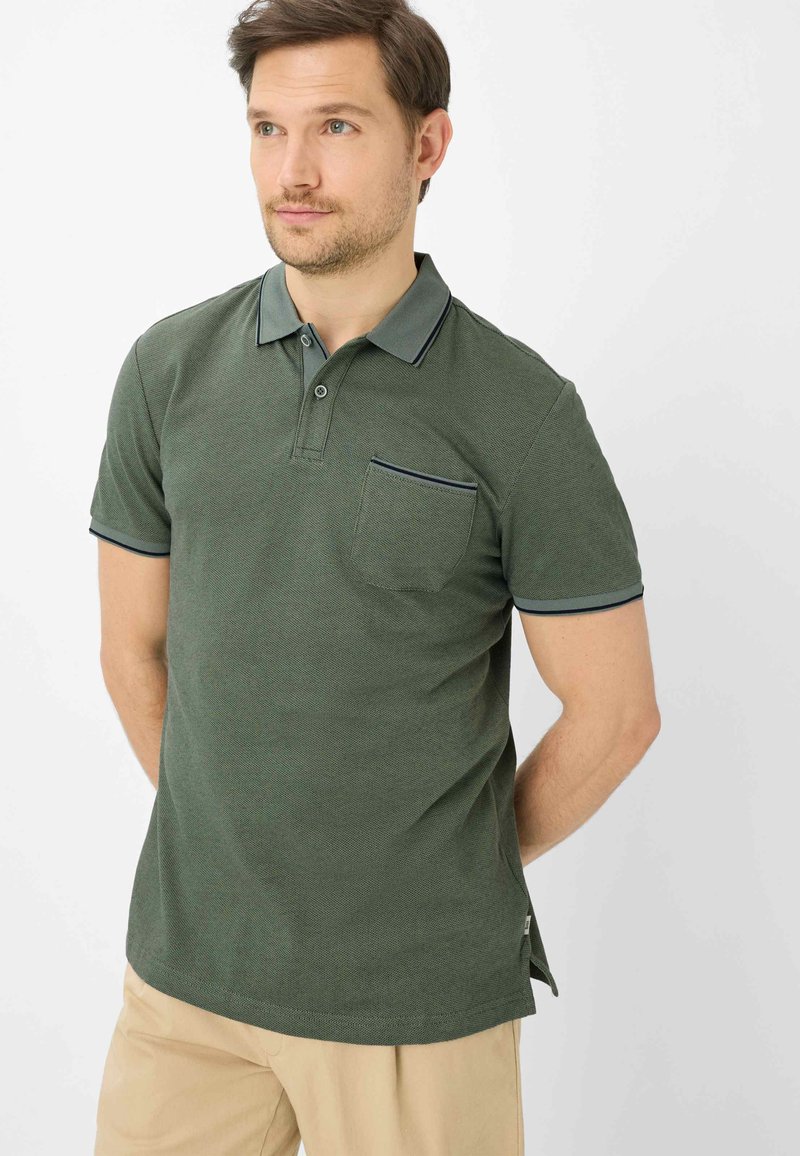 Mann trägt ein dunkelgrünes Polohemd mit kurzen Ärmeln und Brusttasche sowie beige Hose, steht mit den Händen hinter dem Rücken und blickt leicht nach rechts.