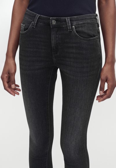 Μαύρο τζιν slim fit που φοριέται από άτομο με τα χέρια χαλαρά στα πλευρά, συνδυασμένο με μαύρο τοπ.