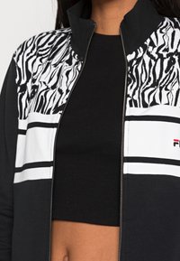 Chaqueta negra con cremallera que presenta un diseño de estampado de cebra en el cuello y franjas horizontales blancas. Tejido suave con un ajuste corto.
