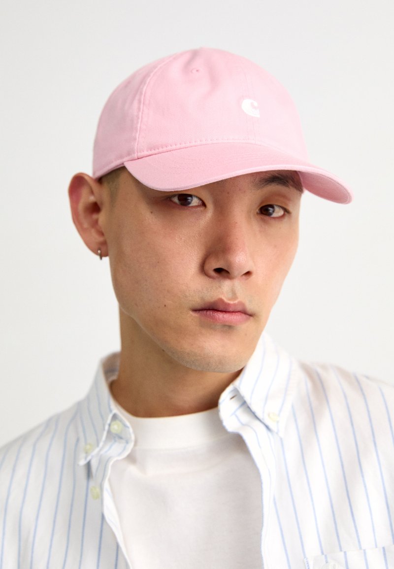 Boné rosa com a aba curva e logótipo branco, usado com uma camisa de botões riscada em azul claro sobre uma t-shirt branca.