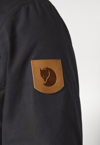 Tamsaus audinio rankovė su ruduo odiniu pleistru, kuriame siuvinėta stilizuota lapės galvos logotipas, pritvirtintas viršutinei rankai.