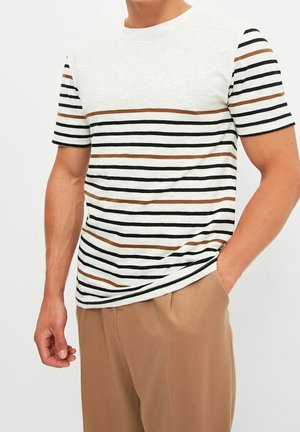 Mand iført hvid kortærmet T-shirt med sorte og brune vandrette striber og beige bukser, den ene hånd i lommen, beskåret ved nakke og lår.