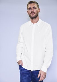 Camicia bianca a maniche lunghe con colletto classico, abbinata a jeans in denim blu con texture. Design semplice e vestibilità rilassata.